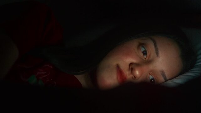 Thoughtful young woman lying in bed at night, scrolling on social media with a soft screen glow on her face, cinematic low light aesthetic.