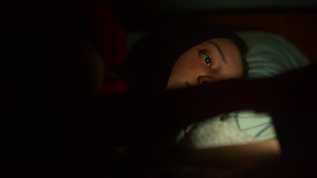 Thoughtful young woman lying in bed at night, scrolling on social media with a soft screen glow on her face, cinematic low light aesthetic.