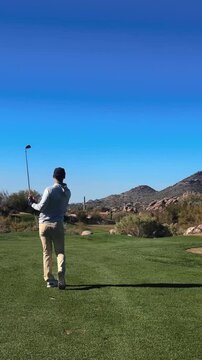 Golfer hits iron with a draw into protected green on pristine Arizona desert golf course, cactus and mountain backdrop, windy, vertical 4k sunny day slow motion