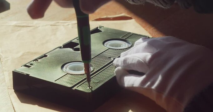 Close-up detail of a repairer using a precision screwdriver to undo screws on a vintage VHS tape. Warm ambient lighting on a wooden desktop.
