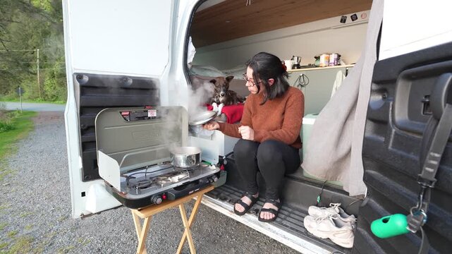 Woman preparing a meal on a camping stove while her dog watches from inside the camper van enjoying van life in the outdoors