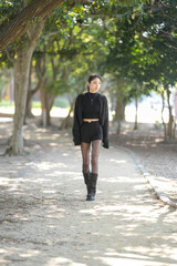 Fototapeta premium Young woman smiles softly, leaning against tree trunk. Black knit top and sparkly cardigan add stylish contrast. Dark shorts and sheer tights complete her trendy look