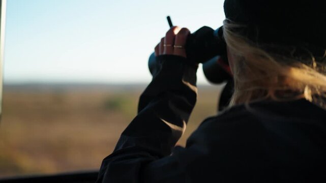 Woman in a vehicle with the wind blowing through her golden hair looking through binoculars into the landscape and horizon which is out of focus