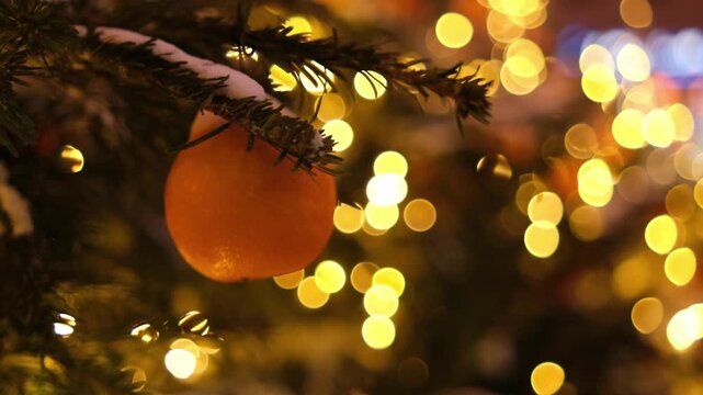 Orange ornament dangles from a snow-dusted pine branch, surrounded by softly glowing Christmas lights, enhancing the holiday spirit