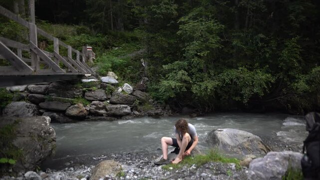 Tired hiker resting by an alpine river in the Swiss forest, dealing with foot blisters after a long hike, remote wilderness, realistic outdoor struggle, raw mountain nature