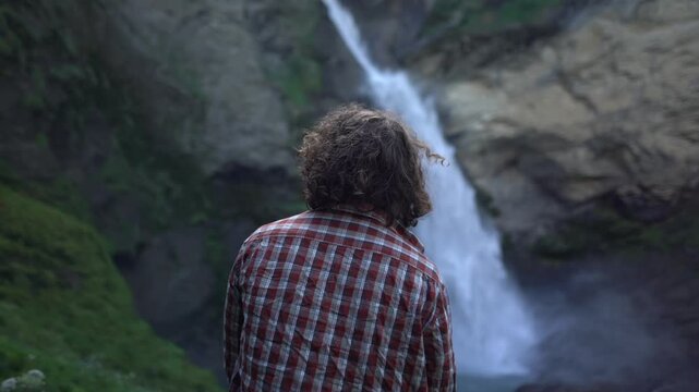 Male hiker standing close to a powerful waterfall in the Swiss Alps, taking a quiet moment in nature. Hiking break at Via Alpina Switzerland Reichenbach waterfall near Meiringen.