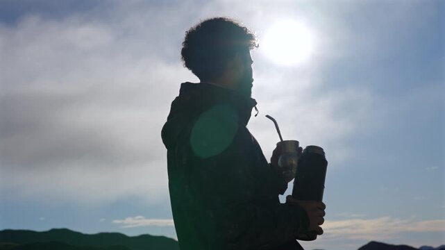 Backlit silhouette of a man drinking yerba mate (Ilex paraguariensis) while holding a thermos outdoors, with strong sunlight and open sky creating a calm contemplative scene