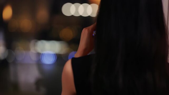 Confident girl swirls red wine in glass, enjoying drinking red pink wine and watches night life of city from top floor of skyscraper, standing by window in dark room. Wine tasting. Holiday party