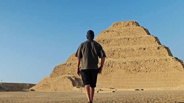 Man walking across the desert plateau of Saqqara as the sun sets, casting a warm, golden glow over the ancient landscape. In the background stands the iconic Step Pyramid of Djoser.