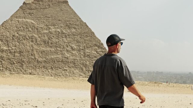 A man is seen walking across the sandy expanse of the Giza Plateau, heading toward the majestic Pyramid of Khafre. The pyramid is clearly distinguished by the surviving limestone casing at its peak.