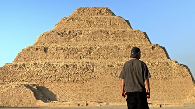 Man walking across the desert plateau of Saqqara as the sun sets, casting a warm, golden glow over the ancient landscape. In the background stands the iconic Step Pyramid of Djoser.