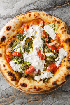 Homemade pizza with stracciatella cheese, pesto, ham and cherry tomatoes close-up on a wooden board on the table. Vertical top view from above
