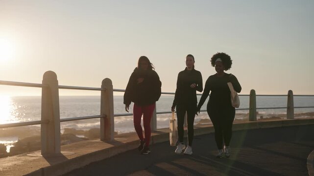 Front view shot of three women walking on a road beside the ocean becoming silhouetted by the sunshine behind them