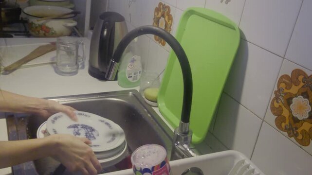 Authentic candid shot of daily household chores showing hands rinsing white patterned dishes under running water in a cozy lived-in kitchen environment perfect for home care and lifestyle branding.