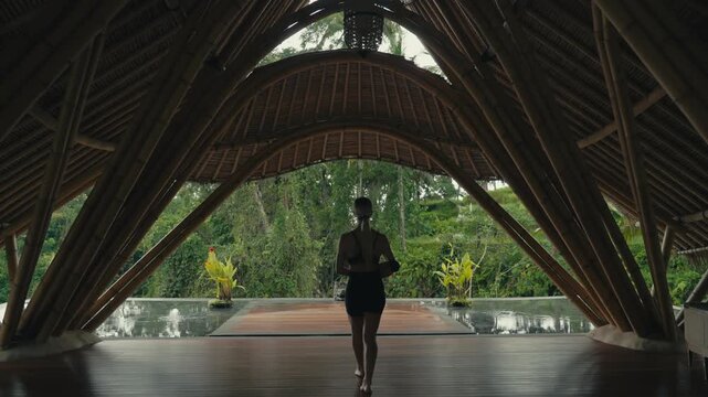 Blonde woman walks through bamboo yoga shala with yoga mat towards outdoor yoga platform