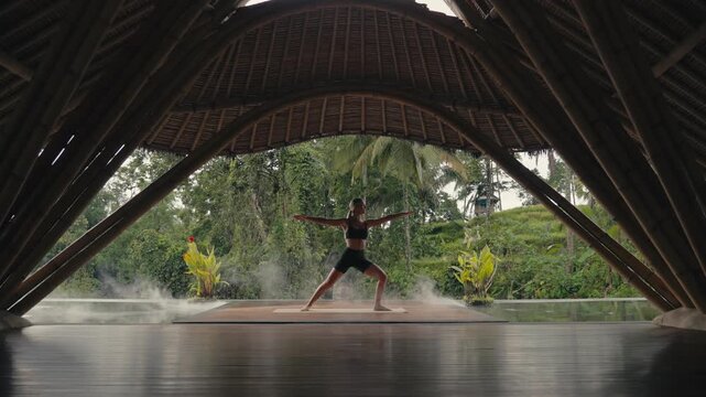 Blonde fit Caucasian Woman Doing Warrior Yoga Pose in tropical Bamboo Yoga Shala with cinematic smoke passing by, cinematic lush green jungle view