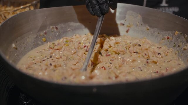 Creamy sauce simmers gently in a pan, stirred with care. Spoon moves through the mixture, blending ingredients with precision. The cook's hands guide the utensil, ensuring even cooking