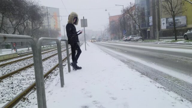 A woman in a dark winter coat stands on a snowy Budapest sidewalk reading a paper document. City traffic passes on Tihamer Utca during a misty winter morning in Hungary in 2026.