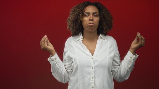 Woman with raised hands pinching fingers in red studio, eyes rolled upward and shoulders relaxed while wearing white shirt and gold ring on finger; frustration.