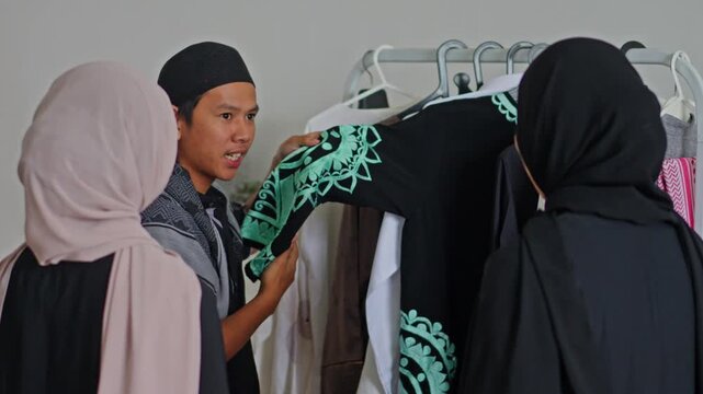 Muslim man shopkeeper showing black abaya with green embroidery to two women at muslim clothes shop