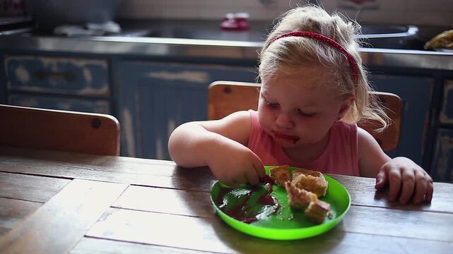 Female toddler eats sandwiches with ketchup at kitchen table at home