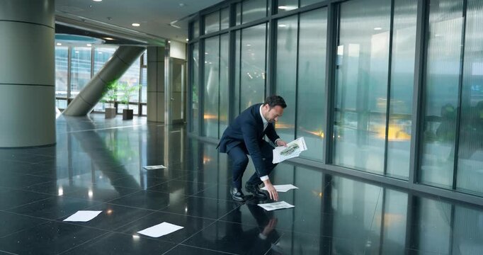 Desperate male manager in blue suit collects scattered report papers from floor after heated conflict with boss in modern corporate office hallway in slow motion