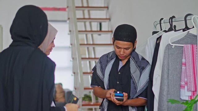 Young man in Muslim attire processing card payment with two women in clothing boutique