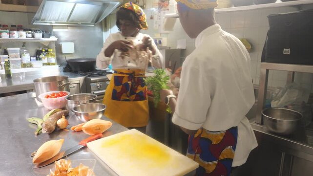 African woman chef washes leafy greens (Amaranthus spp.) beside cassava (Manihot esculenta), plantain (Musa spp.), tomato (Solanum lycopersicum), onion (Allium cepa) and yam (Dioscorea spp.)