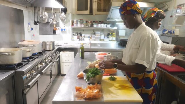 African woman chef sorting leafy greens on a cutting board in a professional kitchen, with large pots on the stove, cassava, yam, plantain, carrot, onion, and tomato prepared on a stainless counter.
