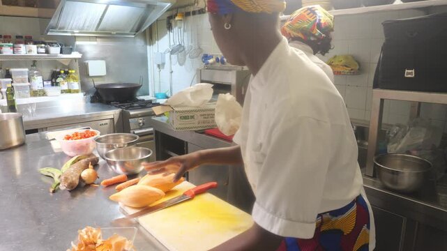 African woman chef cleaning workstation while another washes vegetables in professional kitchen