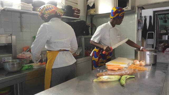 two African women chefs working side by side in a professional kitchen, one chopping vegetables at the counter and the other holding a knife near cassava, plantain, carrot, onion, and a stock pot duri