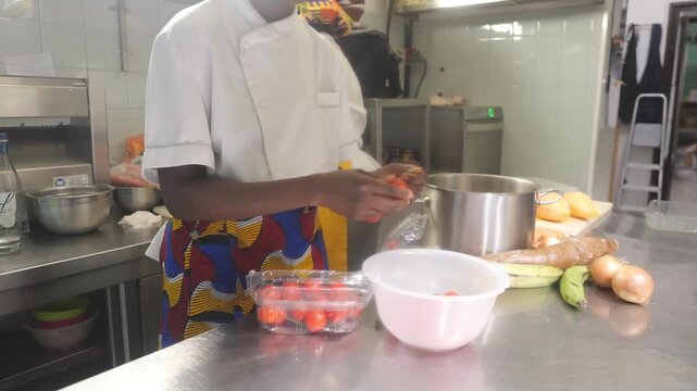 frican woman chef prepares cherry tomato (Solanum lycopersicum) beside a stock pot, with cassava (Manihot esculenta), plantain (Musa spp.), onion (Allium cepa) and yam (Dioscorea spp.) on a kitchen