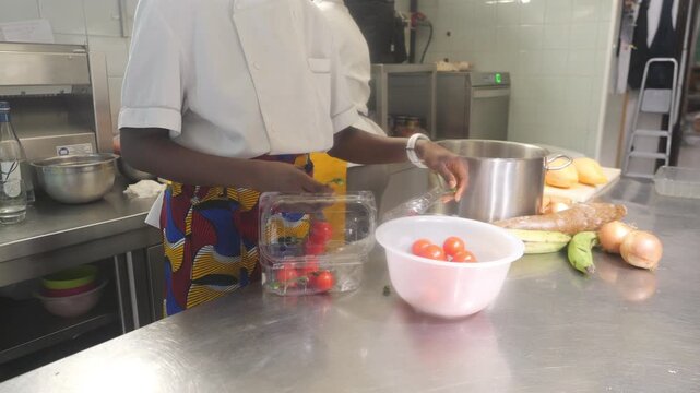 African woman chef preparing ingredients in a professional kitchen, placing cherry tomato into a bowl beside a stainless stock pot, with cassava, plantain, onion, and peeled yam on the counter.