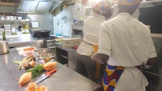 two African women chefs working side by side in a professional kitchen, with cassava mandioca, yam, carrot, onion, tomato, and leafy greens prepared on a stainless counter for a traditional recipe.