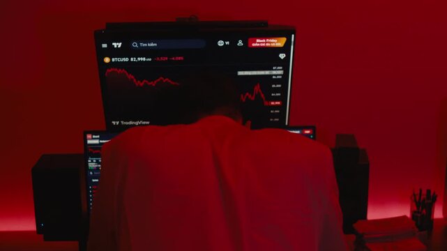 A cinematic shot from behind a trader slumped over his desk in a dark office, with multiple monitors displaying crashing stock market charts in a saturated red light
