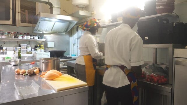 Two African women chefs work in a professional kitchen, with one cleaning the stainless counter while the other washes ingredients at the sink beside cassava (Manihot esculenta) and yam (Dioscorea spp