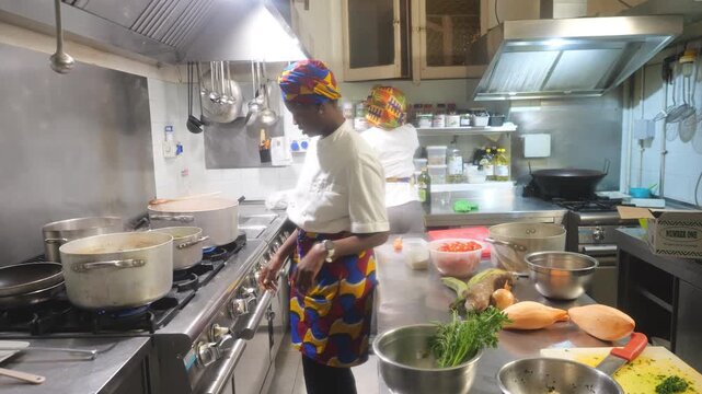 African woman chef prepares leafy greens (Amaranthus spp.) beside cassava (Manihot esculenta), yam (Dioscorea spp.), plantain (Musa spp.), carrot (Daucus carota), onion (Allium cepa) and tomato
