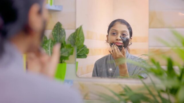 female in bathrobe applying detox charcoal facial mask in front of a bathroom mirror during home skincare routine.