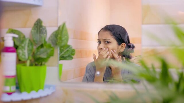 Worried Indian woman touching her cheek and checking pimple in mirror reflection during her skincare routine at home.