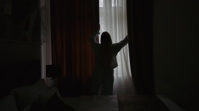 A young woman stands in a bedroom opening the curtains to let in soft morning light.