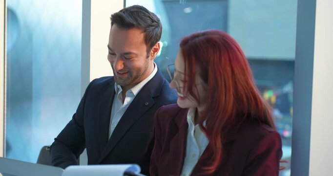 Smiling businessman with laptop and Latina businesswoman with clipboard happily reviewing project documents together leaning on modern office windowsill during golden sunset in slow motion