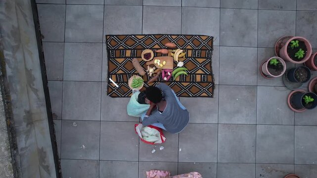 Woman preparing assorted tropical produce on a patterned African fabric, including banana (Musa), cassava (Manihot esculenta), okra (Abelmoschus esculentus) and garlic (Allium sativum), top down.