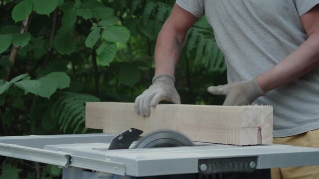 Medium shot of a worker in gloves pushing a large wooden beam through a table saw blade outdoors.