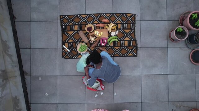 Woman arranging a flat lay of fresh tropical produce on a patterned African textile over tiled patio floor, including plantain cassava, okra, garlic and ginger, with hands styling the scene, top down
