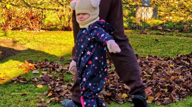 Little child crying while parent raking autumn leaves in home backyard, Small child wearing warm clothes helping rake fallen autumn leaves from green grass
