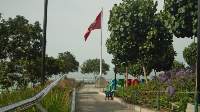 A person walking down a path on a public park called "Parque Bicentenario" with many trees, flowers and plants. A Peruvian flag moves in the wind in the distance. Located in Miraflores, Lima, Peru.