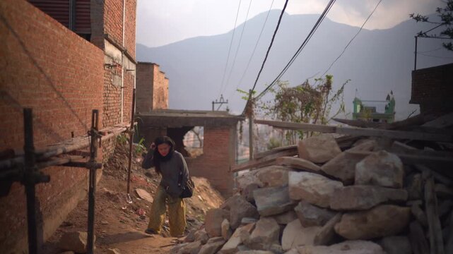 Female traveler walking through damaged residential area in Nepal, brick houses and rubble around, mountains in background, authentic local neighborhood, documentary style travel scene