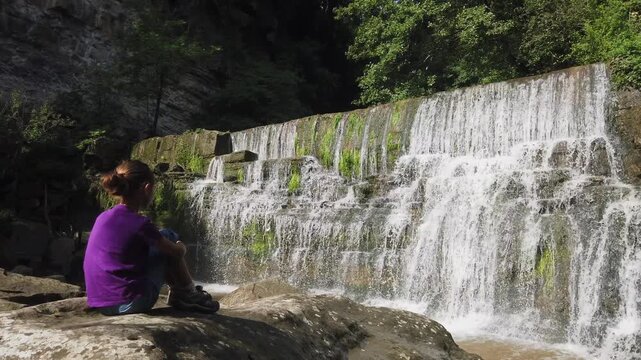 Young girl sitting on a rock looking at the beautiful Salt de Sallent waterfall in Rupit, Catalonia. The water is flowing down the rocks creating a relaxing natural scene