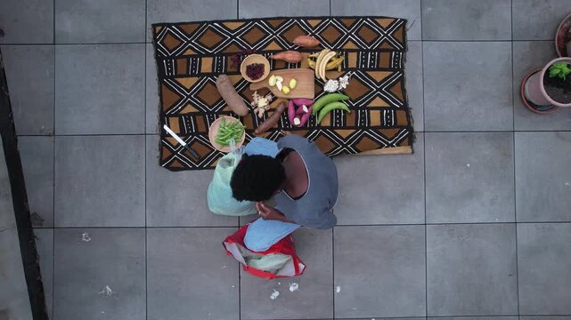Assorted tropical produce including banana (Musa), cassava (Manihot esculenta), okra (Abelmoschus esculentus) and garlic (Allium sativum) arranged on patterned African textile over tile patio floor