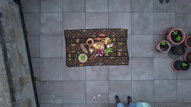 Fresh tropical vegetable and fruit arranged on patterned African textile over tiled patio floor, including banana, cassava, sweet potato, okra and garlic in rustic still life composition, top down